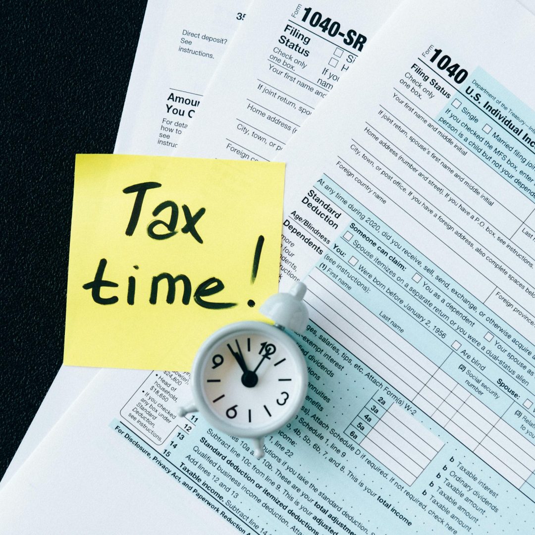 A stack of tax forms with a clock and yellow sticky note saying 'Tax time!' indicating urgency.